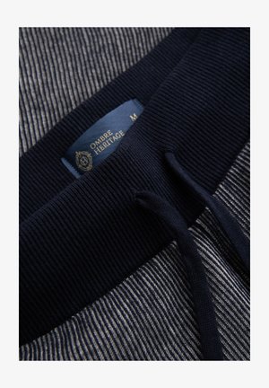 Lähikuva laivastonsinisestä ribbineulosvyötärönauhasta, jossa on vetonarut harmaan ja valkoisen raidallisella kankaalla, ja sininen "Ombre Heritage" -merkkilappu.