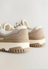 Beige ruskindsneakers med hvide detaljer og en beige gummisål. Har en rund form og tekstureret finish. Lys rosa detalje på hælen.