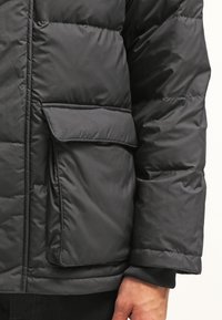 Svart puffjacka med en fyrkantig form, quiltat mönster och en klaffficka på vänster sida. Materialet verkar vara vattenavvisande.