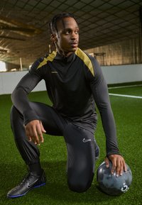 Fekete Nike sportruházatot viselő sportoló, arany szegéllyel, térdel a zöld füvön. Egy szürke labdát tart, bemutatva a karcsú dizájnt és az atletikus illeszkedést.