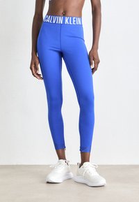 Plave sportske tajice s bijelim pojasom "CALVIN KLEIN", sa elegantnim dizajnom i do gležnja, uparene s bijelim tenisicama.