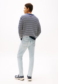 Tânăr stând în picioare, cu fața spre lateral, purtând un pulover în dungi albastre și albe, blugi bleu deschis și adidași navy pe un fundal alb.