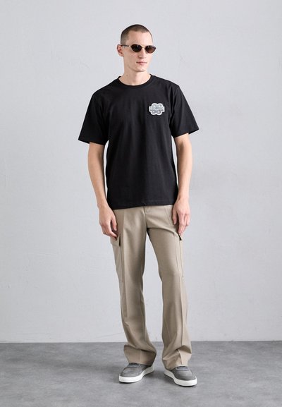 T-shirt en coton noir avec un logo graphique sur la poitrine, associé à un pantalon cargo beige clair et des baskets grises, complété par des lunettes de soleil rondes.