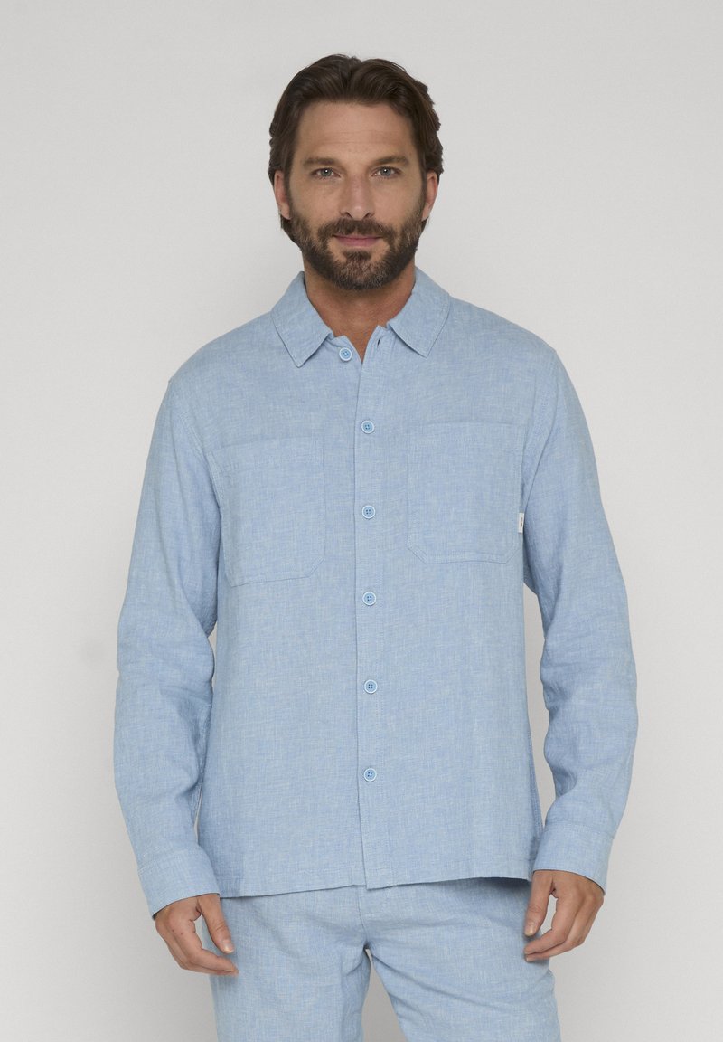 Homme barbu portant une chemise à manches longues bleu clair boutonnée et un pantalon assorti, debout devant un fond blanc uni.
