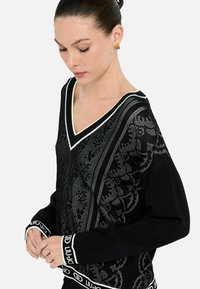 Maglione nero con scollo a V, motivo argentato intricato e rifiniture bianche a contrasto. Presenta una fascia bianca con il marchio sui polsini.