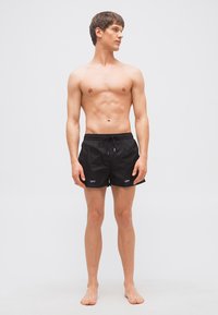 Jeune homme torse nu portant un short de bain noir avec le texte "OFF", debout pieds nus, regardant vers sa droite sur un fond clair uni.