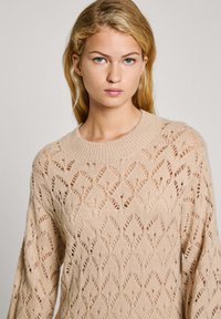 Maglione beige lavorato a maglia con un motivo in pizzo a rombi, scollo a girocollo a coste e maniche lunghe. Finitura testurizzata con dettagli a trama aperta.