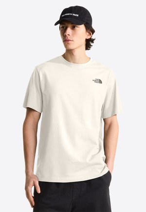 Junger Mann trägt ein hellbeiges The North Face T-Shirt und eine schwarze Kappe, schaut leicht zur Seite mit einer Hand in der Tasche.