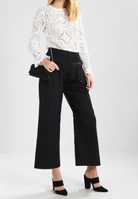 Bílý krajkový top s dlouhými rukávy a květinovými vzory, doplněný o černé kalhoty s vysokým pasem a širokými nohavicemi a černé boty s blokovým podpatkem. Černá psaníčko kabelka.