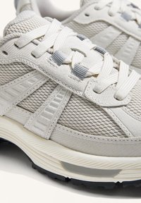 Ljusgrå sportskor med ovandel i mesh, texturerade detaljer, platta skosnören och en svart sulmönstrad sula för grepp.