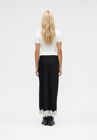 Γυναίκα με μακριά ξανθά μαλλιά που φοράει λευκό τοπ με κοντά μανίκια, μαύρη μακριά φούστα με λευκή δαντέλα στο τελείωμα και μαύρα μπότες με τακούνι, στέκεται με την πλάτη.