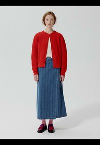 Femme debout portant un cardigan rouge boutonné, un haut blanc, une longue jupe en denim, des chaussettes roses et des chaussures sombres et brillantes avec des pompons.