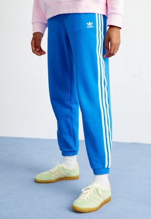 Person wearing blue Adidas track pants with white stripes, light green sneakers, white socks, and a pink sweatshirt standing on blue floor.