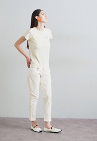 Cremfarbener T-Shirt mit gestickten Akzenten, kombiniert mit hellen Hosen und flachen Schuhen mit einer schwarzen Zehenkappe.