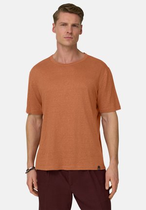 Homme portant un t-shirt à manches courtes de couleur rouille et un pantalon sombre, debout devant un fond clair uni.