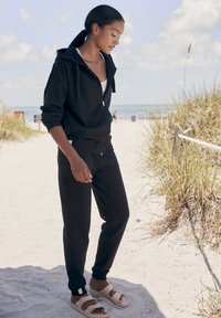 Sudadera negra con cremallera y corte relajado, combinada con pantalones deportivos negros a juego. Sandalias de tiras anchas en color beige claro. Fondo de playa.