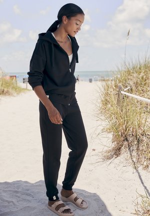 Black zip-up hoodie with a relaxed fit, paired with coordinating black sweatpants. Sandals with a wide strap and light tan color. Beach background.