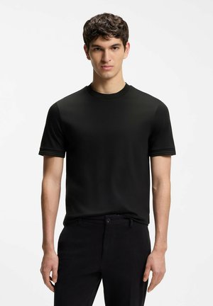 Junger Mann mit kurzen dunklen Haaren, der ein schlichtes schwarzes kurzärmeliges T-Shirt und schwarze Hosen trägt, vor einem weißen Hintergrund stehend.