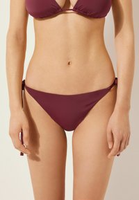 Burgunderfärgad bikiniunderdel med justerbara sidband, med en minimalistisk design och mjuk textur, modellerad på en neutral bakgrund.