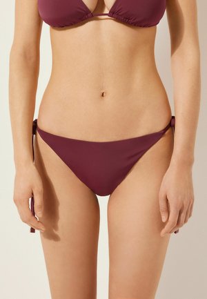 Bas de bikini bourgogne avec liens latéraux ajustables, présentant un design minimal et une texture lisse, mis en valeur sur un fond neutre.
