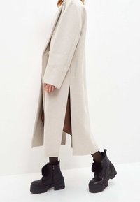 Abrigo largo beige con doble botonadura, slits laterales, usado sobre leggings negros texturizados y botas negras gruesas hasta el tobillo con acentos de piel.