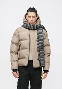 Μπεζ μπουφάν τύπου puffer με υφή, τσέπες με φερμουάρ και ψηλό γιακά, σε συνδυασμό με ριγέ μαύρο και γκρι κασκόλ. Μαύροι cargo παντελόνια.