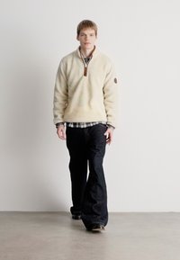 Lys beige fleece pullover med lynlås krave og brune læderdetaljer, parret med mørk denim jeans og en ternet skjorte underneath.