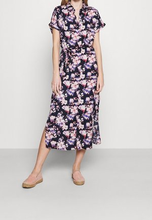 Frau trägt ein marineblaues Midikleid mit Blumenmuster, Taillengürtel, kurzen Ärmeln und rosa Slippern, steht vor einem schlichten hellen Hintergrund.