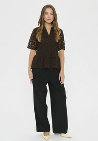 Pikkade juustega naine kannab lühikeste varrukatega tumeda pruunist pitsist pluusi, musti laia säärega pükse ja beeži teravatesse kingadesse, seistes valge tausta ees.