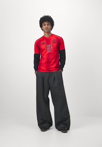 Rød Adidas-trøye med svarte lange ermer, med logo og strukturert mønster, parret med mørke vide bukser og svarte sko.