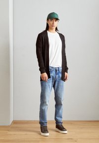 Černý pletený cardigan přes bílé tričko, v kombinaci s modrými džíny a hnědými teniskami. Modelka má na sobě zelenou baseballovou čepici a stojí na dřevěné podlaze.