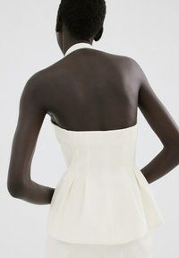 Persona de piel oscura con cabello corto, vistiendo un top blanco sin tirantes y plisado, vista desde atrás contra un fondo liso y claro.