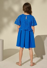 Blauwe jurk met kanten bovenkant en peplum taille, korte mouwen en een uitlopende rok. Detail met sleuf achter. Ggedragen met zilveren sandalen.