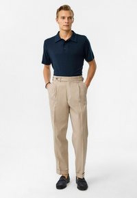 Marineblauwe poloshirt met een knoopsluiting op de kraag, gecombineerd met hooggesneden beige broeken met voorplooien en schuine zijzakken. Zwarte instappers.