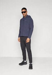 Άντρας με σκούρο ναυτικό μπουφάν με κουκούλα, μαύρο tapered παντελόνι, μαύρα αθλητικά παπούτσια, λευκές κάλτσες και μεγάλα μαύρα γυαλιά ηλίου, στέκεται μπροστά από απλή φόντο.