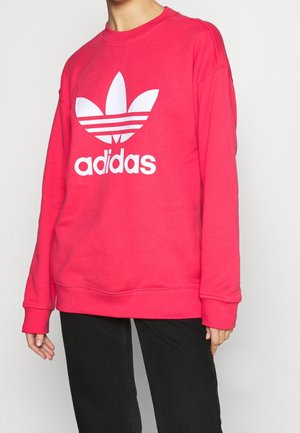 Osoba nosící jasně růžovou mikinu Adidas s bílým logem trefoilu a černé kalhoty na jednoduchém světlém pozadí.