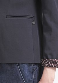 Námořnický blejzr s hladkou texturou, který má boční kapsu a dekorativní knoflík. Manžety jsou zdobeny hnědým vzorovaným lemem.