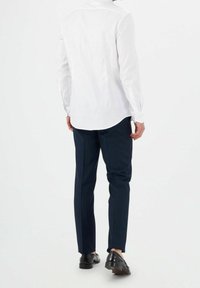 Bílá košile s dlouhým rukávem a zaobleným lemem, ve spojení s námořnicky modrými kalhotami s proužky a černými botami, viděná zezadu.