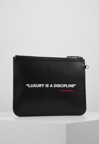 Černé kožené psaníčko s hladkým povrchem má zapínání na zip. Text v bílé a červené barvě hlásá: "LUXUS JE DISCIPLÍNA" - KARL LAGERFELD.