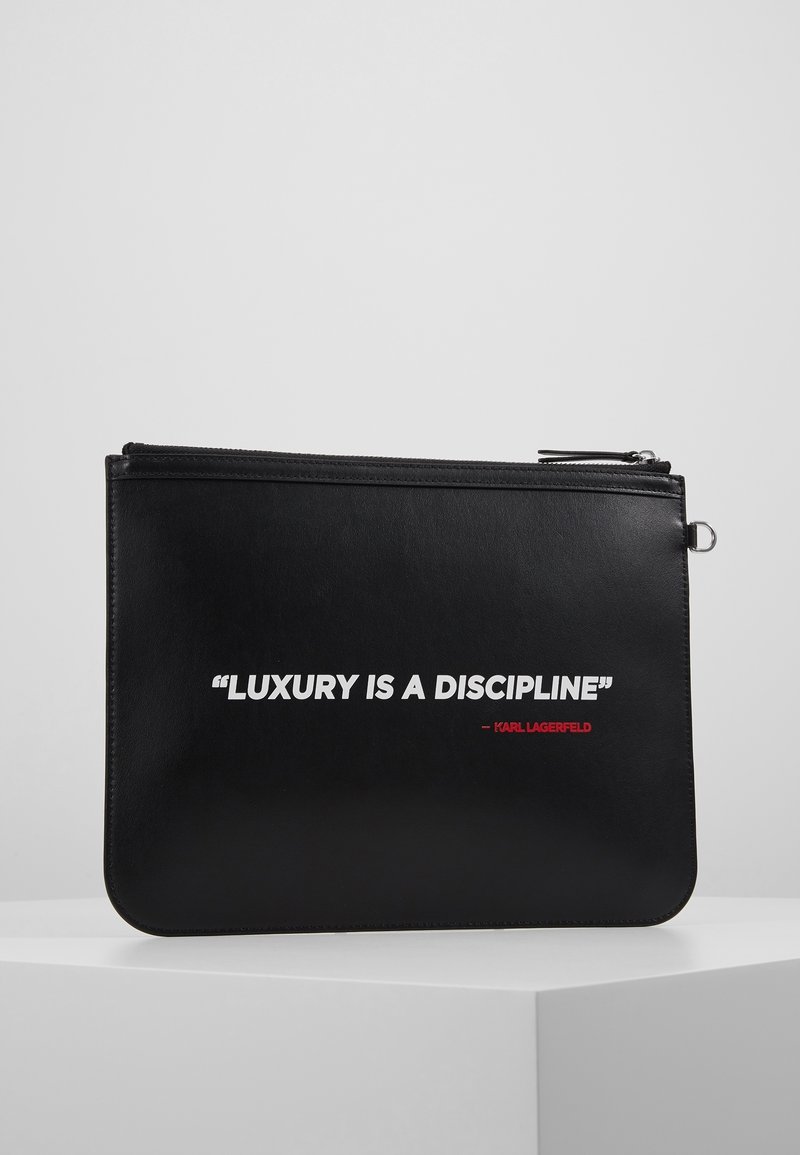 Černé kožené psaníčko s hladkým povrchem má zapínání na zip. Text v bílé a červené barvě hlásá: "LUXUS JE DISCIPLÍNA" - KARL LAGERFELD.