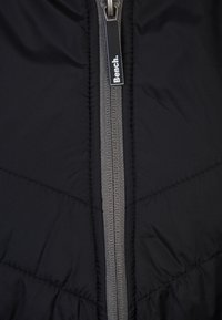 Veste matelassée noire à texture lisse, avec une fermeture éclair centrale grise, et une petite étiquette blanche portant l'inscription "Bench." sur le curseur de la fermeture éclair.