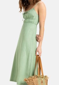 Robe en lin vert clair avec un corsage ajusté, des bretelles réglables et une jupe évasée. Associée à un sac fourre-tout tissé beige avec des accents verts.