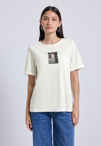 Camiseta de crema clara con mangas cortas, escote redondo, con un diseño impreso en negro y rosa en el centro. Textura de tejido suave.