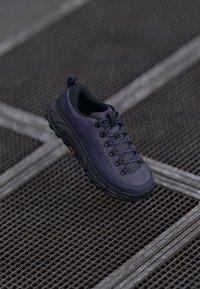 Zapatilla de senderismo morada con un upper texturizado, diseño con cordones, talón acolchado y una suela negra resistente, situada sobre un fondo de rejilla metálica.