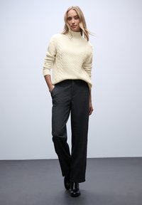 Kremowy sweter w warkocze z wysokim kołnierzem, zestawiony z ciemnoszarymi spodniami o szerokich nogawkach i czarnymi botkami za kostkę, na neutralnym tle.