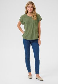Top verde oliva a maniche corte con motivo ad occhiello, abbinato a jeans blu aderenti. Ballerine beige chiaro completano l'outfit.
