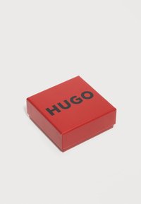 Κόκκινο τετράγωνο κουτί με μαύρο λογότυπο «HUGO», τοποθετημένο σε απλό λευκό φόντο.