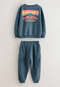 Felpa blu con grafica di montagna e testo "Superdry Outdoor", abbinata a pantaloni jogger cargo blu coordinati.