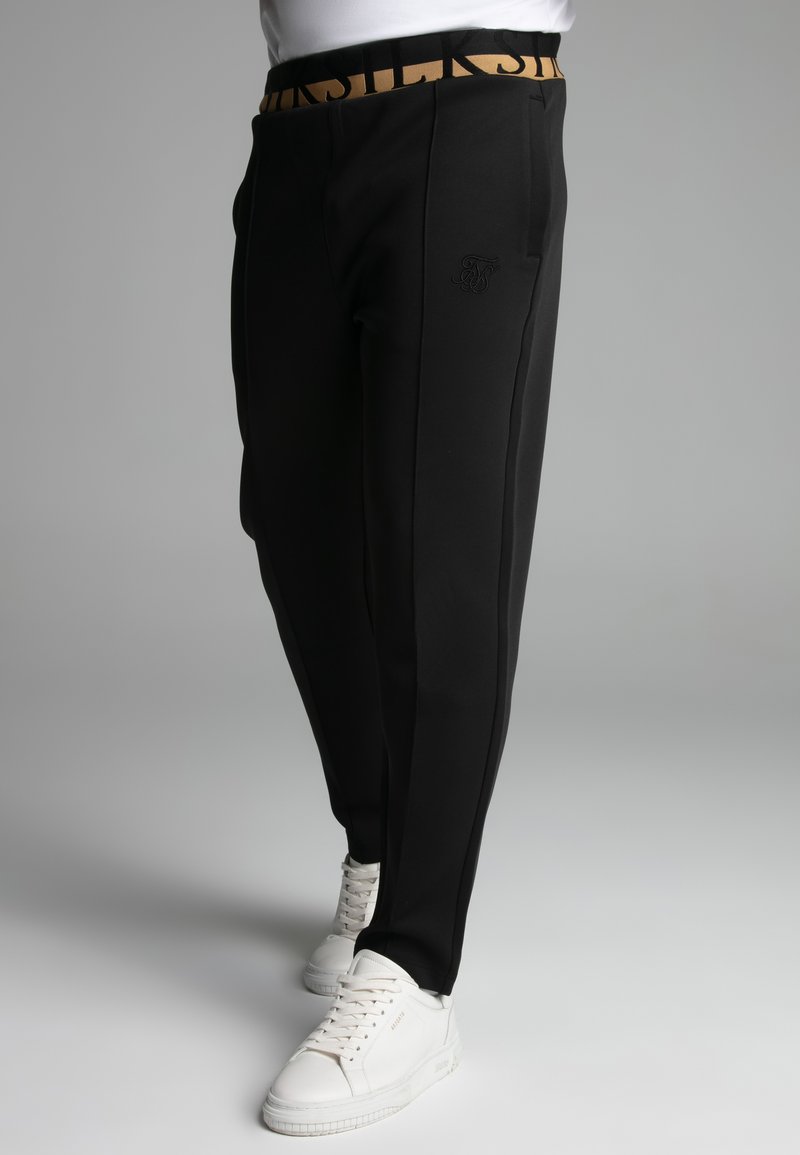 Zwarte sportbroek met een gouden merkband, voorzien van zakken en een gestructureerde afwerking. Witte sneakers maken de outfit compleet.