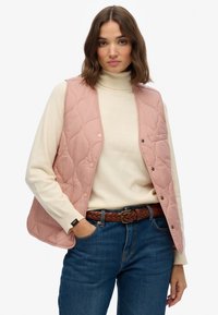 Gilet matelassé rose avec des boutons-pression, porté sur un col roulé crème. Présente une ceinture tissée marron et un jean bleu avec des poches avant.